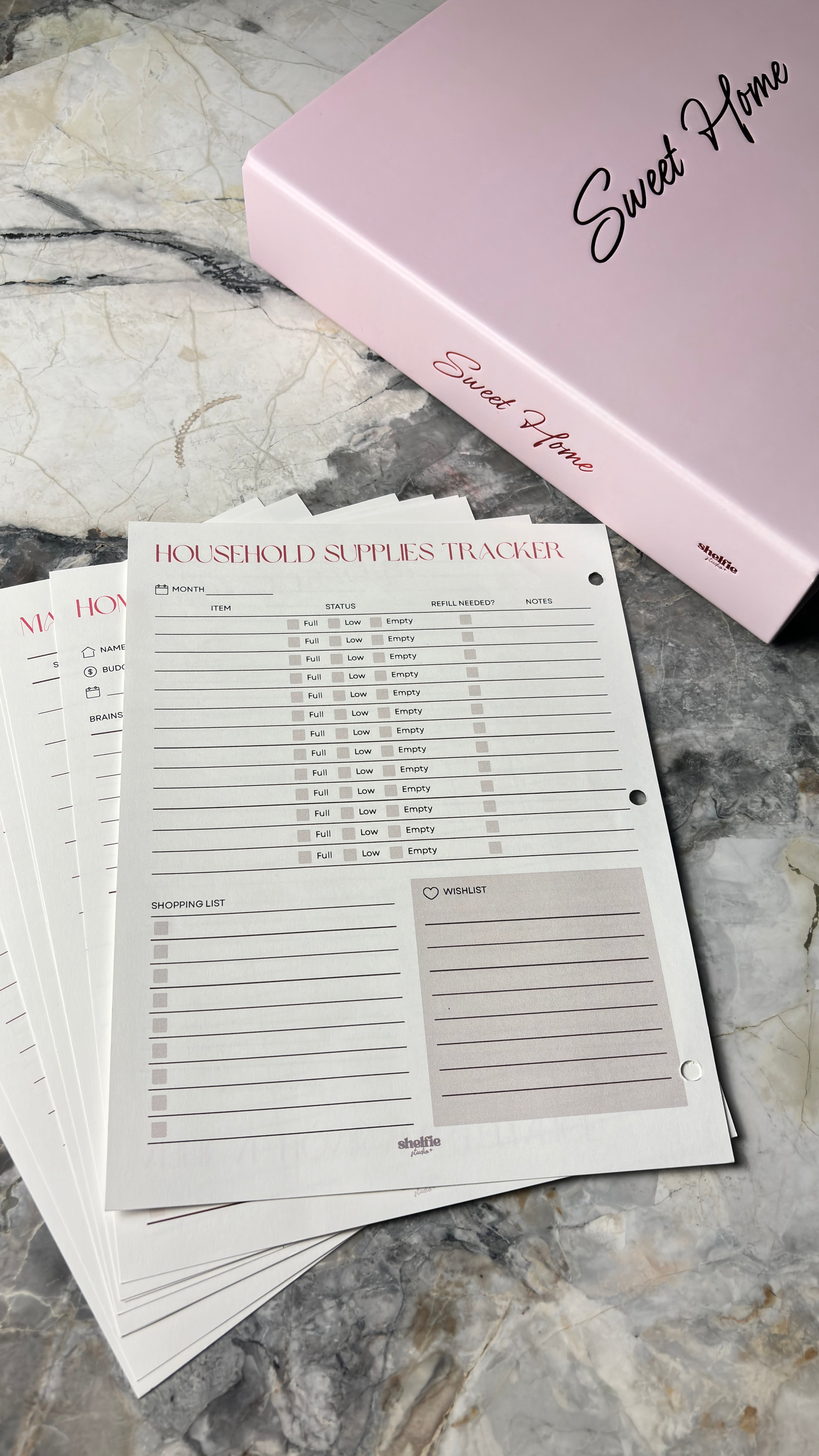 Household supplies tracker sheets with a pink binder labeled 'Sweet Home' on a marble surface.